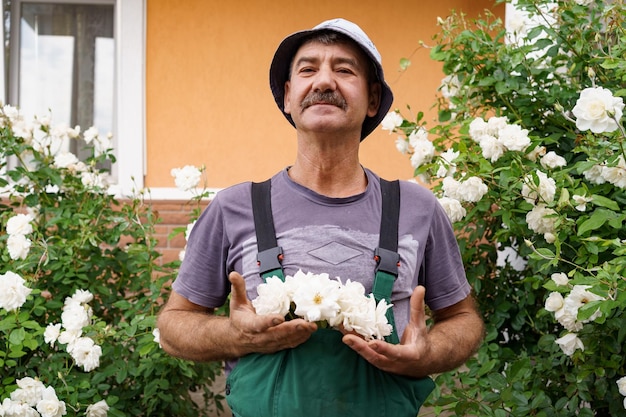 Lächelndes Mannporträt mit einer weißen Rose auf seinem Hut mit Rosenbüschen im Hintergrund