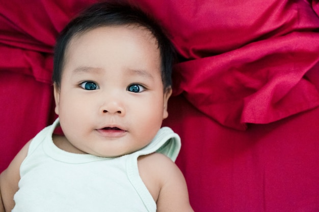 Lächelndes nettes Babykind, das auf rotem Bett, Fokus auf dem Gesicht liegt und Hand wellenartig bewegen