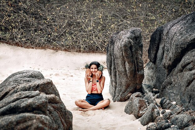 Lächelndes nettes Mädchen in der Lotoshaltung am Strand hält zwei Ananas; lustiges Yoga-Konzept