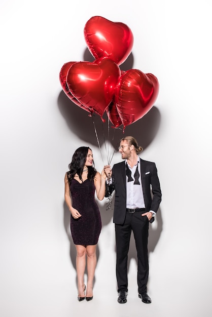 lächelndes Paar hält rote herzförmige Ballons am Valentinstag auf weiß