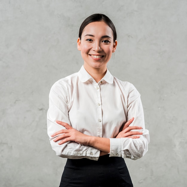 Foto lächelndes porträt einer asiatischen jungen frau mit ihren armen kreuzte das schauen zur kamera gegen graue betonmauer