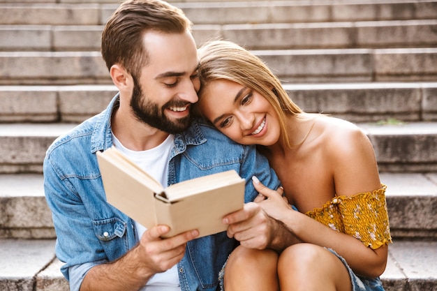 Lächelndes schönes junges Paar, das zusammen auf der Treppe sitzt, ein Buch liest, sich umarmt