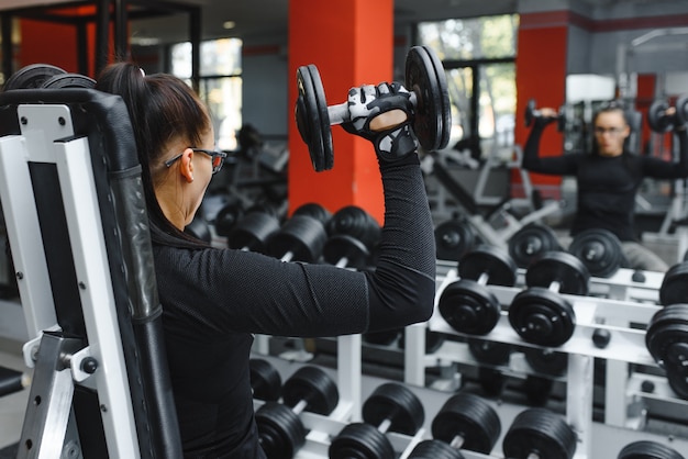 Lächelndes schönes Mädchen mit schweren Hanteln vor dem Spiegel in einer Sporthalle. Starke junge schöne Frau ist im Fitnessstudio mit Hanteln vor einem Spiegel beschäftigt.