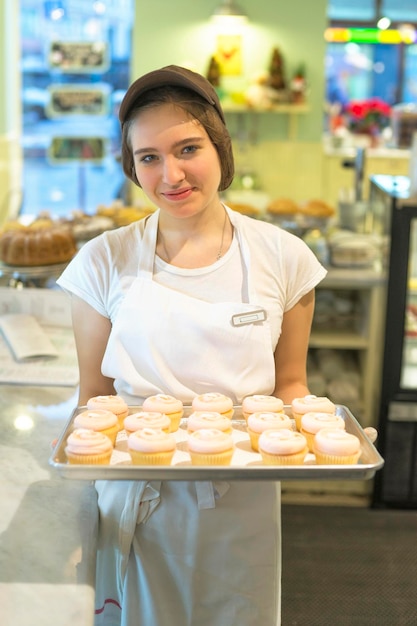 Lächelndes schüchternes Mädchen in der Mütze mit Cupcakes am Tablett in der Bäckerei