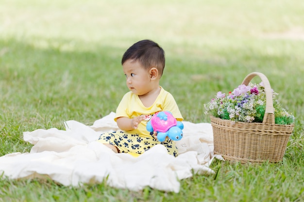 Lächelndes schwarzes Haar des asiatischen Jungen und Augenkleinkind sitzen auf weißer Baumwolle im grünen Gras allein und spielen Spielzeug.