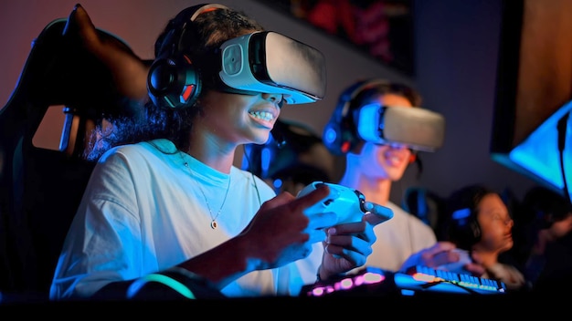 Lächelndes schwarzes Teenager-Mädchen im VR-Headset spielt Videospiele mit Gamepad im Videospielclub