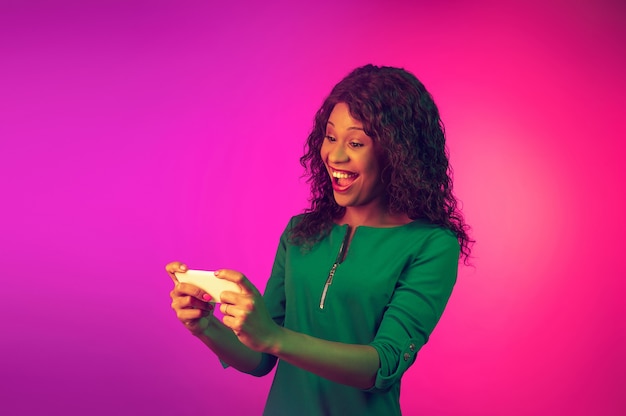 Lächelndes, scrollendes Telefon. Afroamerikanische junge Frau auf rosa Hintergrund mit Farbverlauf im Neonlicht. Schönes weibliches Modell. Konzept der menschlichen Emotionen, Gesichtsausdruck, Verkauf, Anzeige. Flyer mit Exemplar.