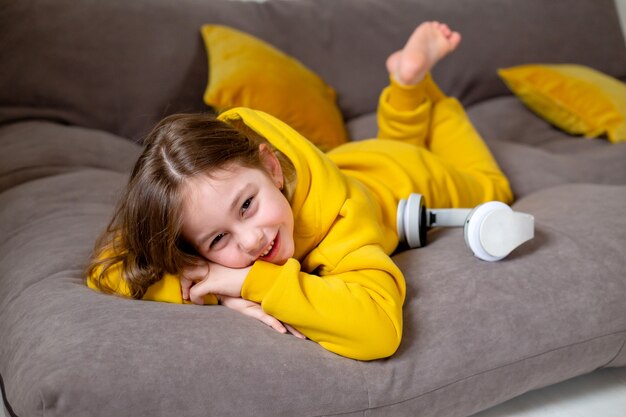 Lächelndes süßes kleines Mädchen in gelber Kleidung im Bett liegend mit Kopfhörern beim Musikhören