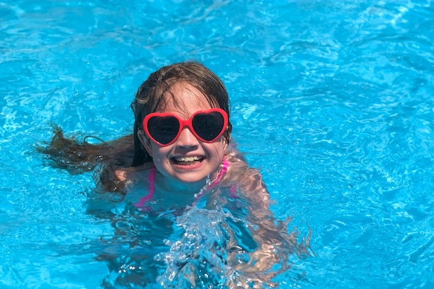 Lächelndes süßes kleines Mädchen mit herzförmiger Sonnenbrille im Schwimmbad an einem sonnigen Tag