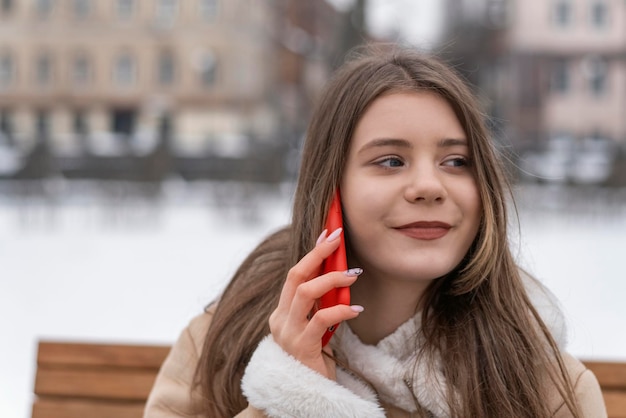 Lächelndes süßes Mädchen sitzt auf einer Parkbank und spricht auf dem Smartphone Lächelnde junge Frau in beigem Schaffellmantel ruht draußen