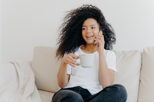 Lächelndes tausendjähriges Mädchen mit Afro-Haaren, genießt Handygespräche mit heißem Getränk in der Tasse, hat gesunde Haut, ist lässig gekleidet, sitzt auf einem weißen Sofa zu Hause in einer modernen Wohnung und bestellt die Lieferung