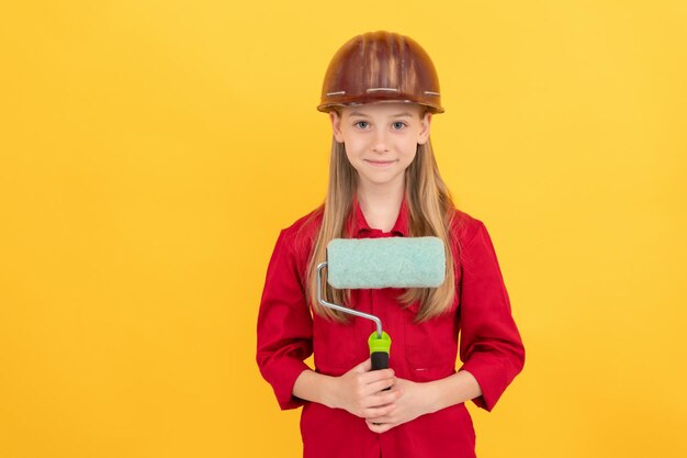 Lächelndes Teenager-Kind im Bauarbeiterhelm mit Farbroller an gelber Wand