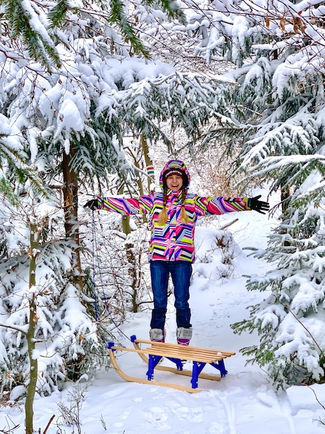 Lächelndes Teenager-Mädchen mit Schlitten, das durch einen schneebedeckten Wald läuft. Glückliches Kind und wunderschöne Winterlandschaft