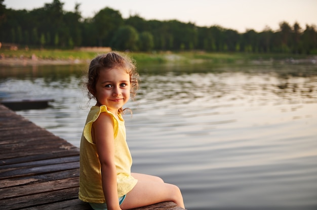 Lächelndes und glückliches Baby, das am Sommerabend auf einem Pier an einem See sitzt. Schönheit in der Natur, Sommerthemen, laue Sommerabende