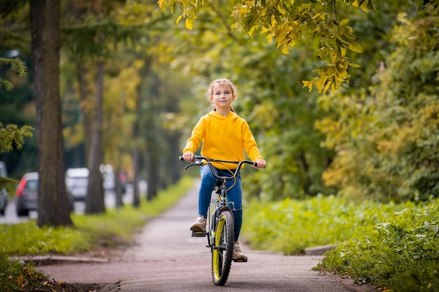 Lächelndes zehnjähriges Mädchen in einem gelben Pullover fährt mit dem Fahrrad auf der Herbststraße unter den Zweigen eines roten Ebereschenbaums