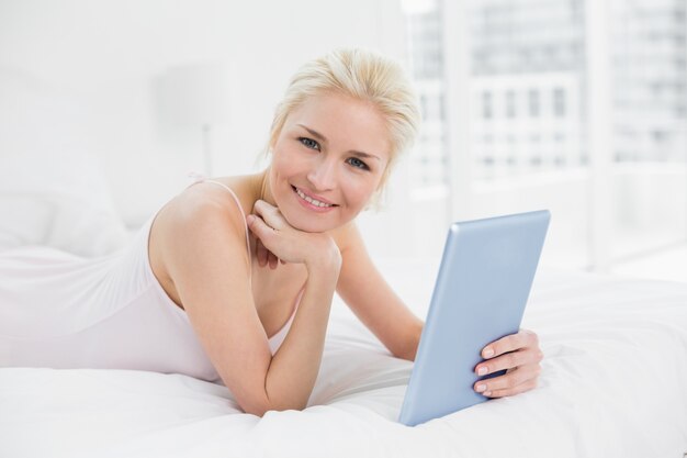 Lächelndes zufälliges junges blondes mit Tablet-PC im Bett