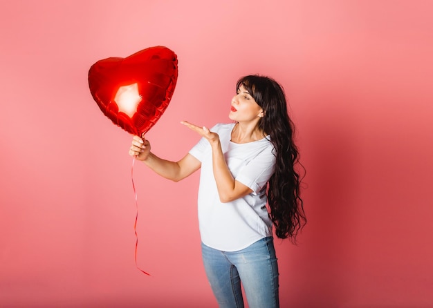 Lächelndes zufriedenes kaukasisches Mädchen, das mit einem roten herzförmigen Ballon auf einem rosa Hintergrund posiert. Valentinstag