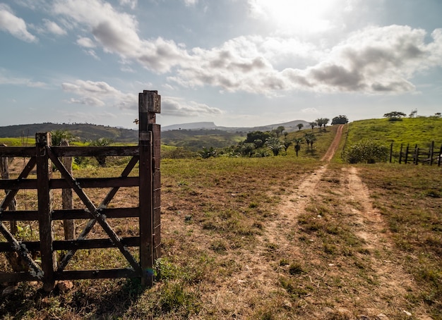 Ländliches Bild auf einem Bauernhof mit Holztor, Feldweg, Sonnenschein und Hügeln im Hintergrund.