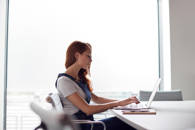 Lässig gekleidete junge Geschäftsfrau, die am Schreibtisch am modernen Arbeitsplatz am Laptop arbeitet