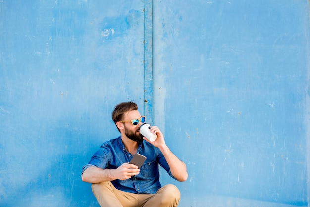 Lässig gekleideter Mann in blauem Hemd, der mit Telefon und Kaffee zum Mitnehmen auf dem blauen Wandhintergrund sitzt. Breite Aufnahme mit Kopierbereich