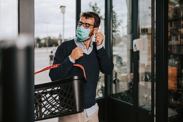 Lässig gekleideter mann mittleren alters mit gesichtsschutzmaske und handschuhen beim betreten des supermarkts oder lebensmittelgeschäfts. pandemie- oder epidemie-lifestyle- und konsumkonzept.