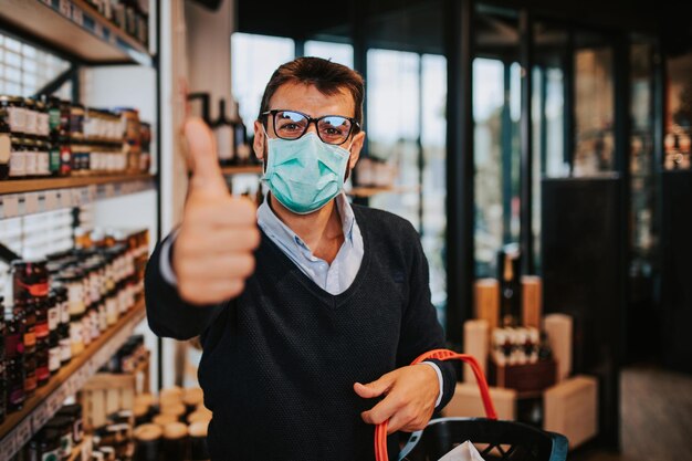 Lässig gekleideter Mann mittleren Alters mit Gesichtsschutzmaske und Handschuhen, der gesunde Lebensmittel und Getränke in einem modernen Supermarkt oder Lebensmittelgeschäft kauft. Pandemie oder epidemisches Lebensstil- und Konsumkonzept.