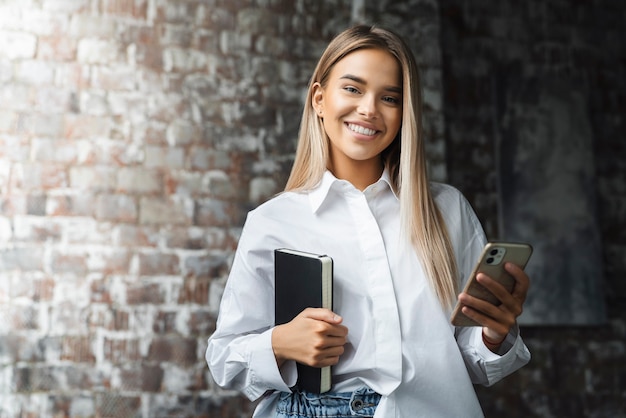 Lässig gekleidetes Mädchen, das ein Notizbuch hält und ein Handy für die Arbeit benutzt.