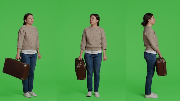 Lässige Person mit Vintage-Koffer im Studio, posiert mit Broefcase vor Ganzkörper-Greenscreen-Hintergrund. Erwachsene Frau hält Reisegepäck vor der Kamera und steht vor einem isolierten grünen Bildschirm.