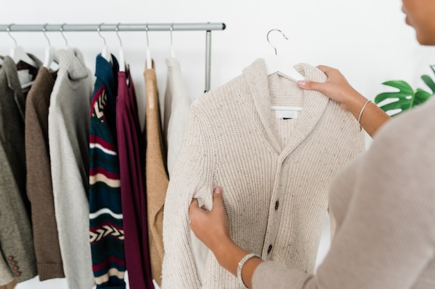 Lässige weiße Strickjacke auf Kleiderbügel, gehalten von einem jungen Shopaholic, der neue warme Kleidung für den Herbst auswählt