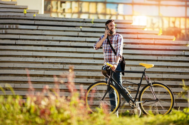 Lässiger Geschäftsmann, der mit dem Fahrrad zur Arbeit geht. Er schiebt Fahrrad und benutzt Telefon.