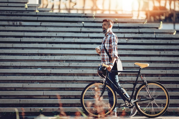 Lässiger, gutaussehender Geschäftsmann, der mit dem Fahrrad zur Arbeit geht. Er schiebt sein Fahrrad und trinkt Kaffee zum Mitnehmen.