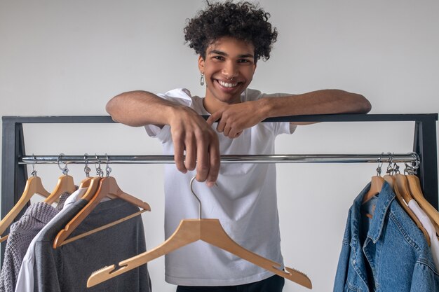 Lässiger Kleidungsstil. . Afroamerikaner junger Mann in der Nähe von Kleiderbügeln mit lächelnden Kleidern