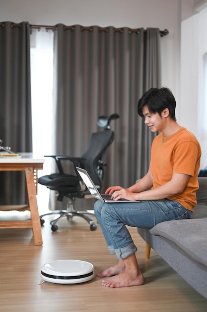 Lässiger Mann mit Laptop auf dem Sofa und Staubsaugerroboter im Wohnzimmer