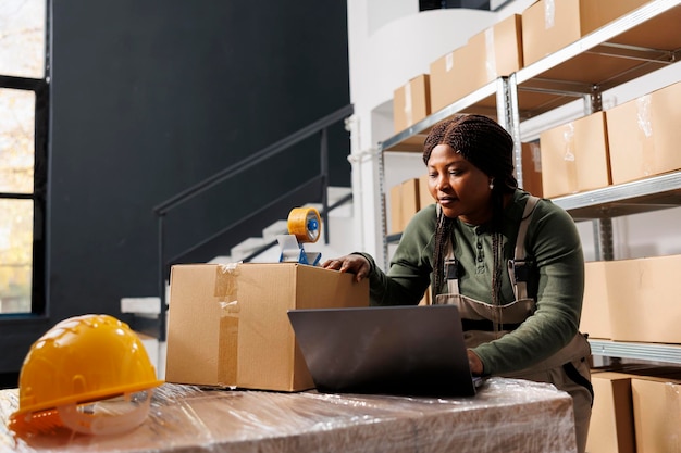 Lagerarbeiter analysiert den Warenbestandsbericht auf einem Laptop, bevor er mit der Arbeit an Kundenbestellungen im Lagerraum beginnt. Afroamerikanischer Manager trägt Industrieoverall im Lager