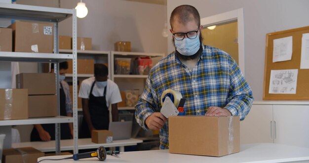 Lagerarbeiter mit Maske verpackt Karton mit Klebeband