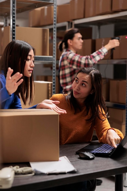 Lagerbetreiber beaufsichtigt Mitarbeiter, der auf einem Laptop in der Lagerhalle Inventar erstellt. Junge asiatische Mitarbeiter diskutieren über die Logistik und den Vertrieb von Waren.