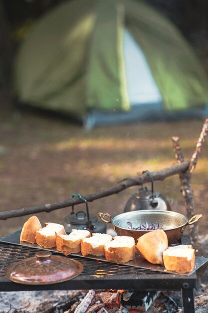 Lagerfeuer und Kochpfanne sind im Vordergrund und fokussiert, es gibt ein Zelt im Hintergrund und defokussiert