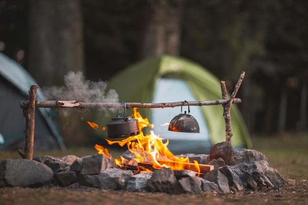 Lagerfeuer und Teekanne sind im Vordergrund und fokussiert, im Hintergrund befindet sich ein Zelt und ist defokussiert