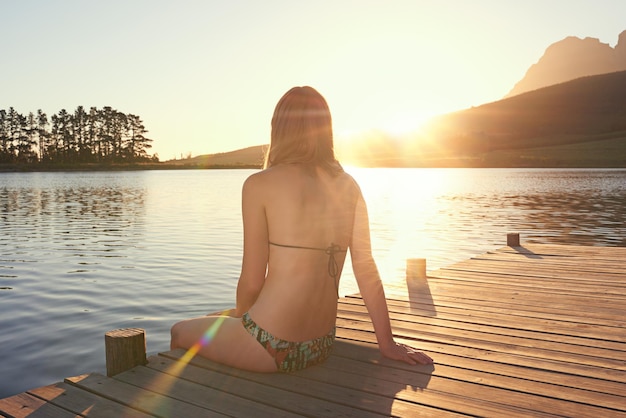 El lago al atardecer Foto de una mujer joven en bikini sentada en un muelle junto a un lago al atardecer