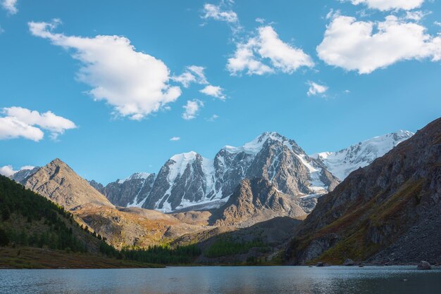 Lago alpino de color azul puro con vista a la colina del bosque y montañas gigantes de nieve en un día soleado de otoño Lago glacial contra una enorme cadena montañosa cubierta de nieve bajo un sol brillante Colores vivos de otoño en altas montañas