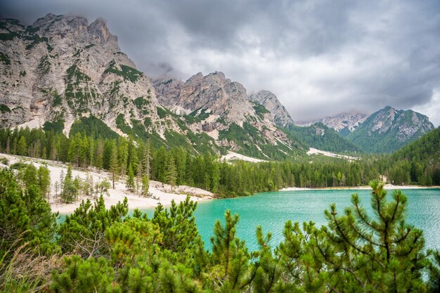 Foto lago braies cercado por florestas de pinheiros e as cordilheiras rochosas das dolomitas em um dia nublado na itália