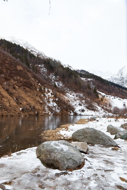 Lago da montanha do inverno na neve no fundo da lagoa da paisagem da montanha com gelo no primeiro plano das pedras da água