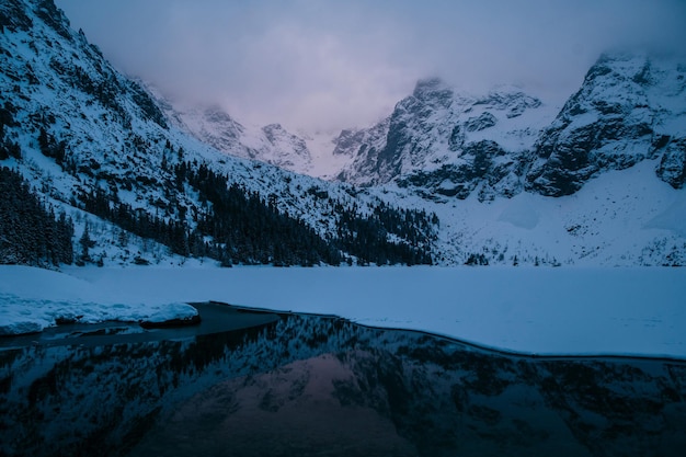 Un lago se encuentra en medio de las laderas cubiertas de nieve de una montaña rodeada por un pintoresco paisaje natural con nubes flotando en el cielo sobre Morskie Oko Polonia