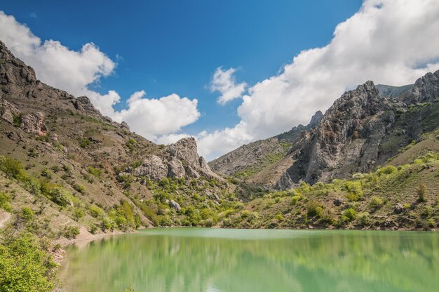 Lago de montaña con agua verde brillante, piedra grande en primer plano en la orilla. Árboles y plantas verdes en segundo plano.