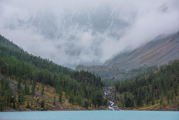 Lago de montaña azul y colinas forestales con cimas de abeto puntiagudas contra grandes montañas nevadas en espesas nubes bajas El arroyo de montaña fluye hacia el lago glacial Cimas de abetos pico y alta cordillera de nieve en densa niebla