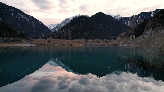 Lago de montaña Issyk con agua de espejo al atardecer. Se reflejan las nubes, los árboles y las montañas.