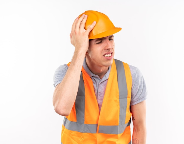 Foto lamentado con los ojos cerrados joven constructor en uniforme poniendo la mano en la cabeza aislada en la pared blanca