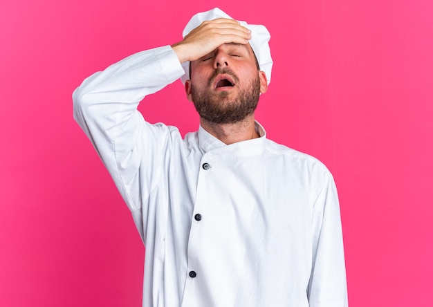 Lamentando a joven cocinero de sexo masculino caucásico en uniforme de chef y gorra manteniendo la mano en la frente con los ojos cerrados aislados en la pared rosa