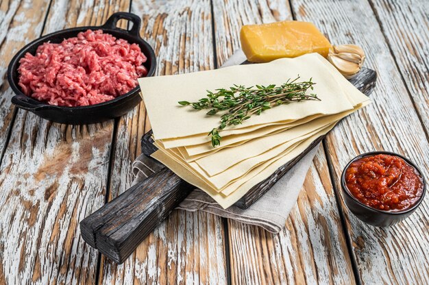 Láminas crudas de lasaña y otros ingredientes. Fondo de madera blanca. Vista superior.