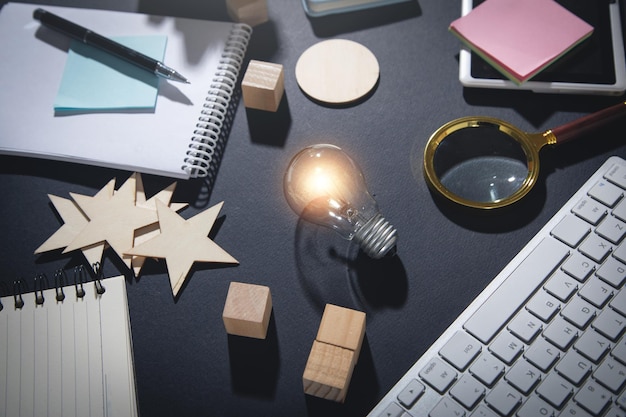 Lâmpada, teclado de computador, cubos de madeira, lupa, estrelas de madeira, caneta, bloco de notas em cima da mesa.